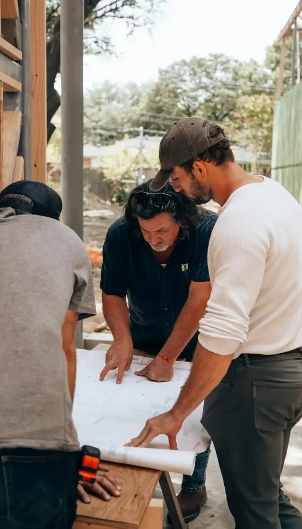 Drei Männer beugen sich draußen über einen ausgerollten Plan, neben einem Holzständerwerk.