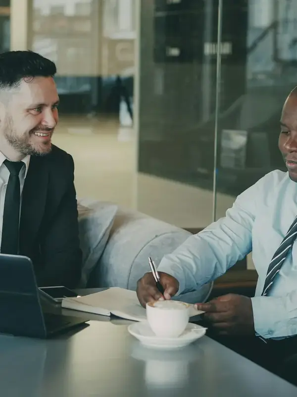 Zwei Männer lächeln bei einem Kaffee, auf dem Tisch ein Computer und Unterlagen.