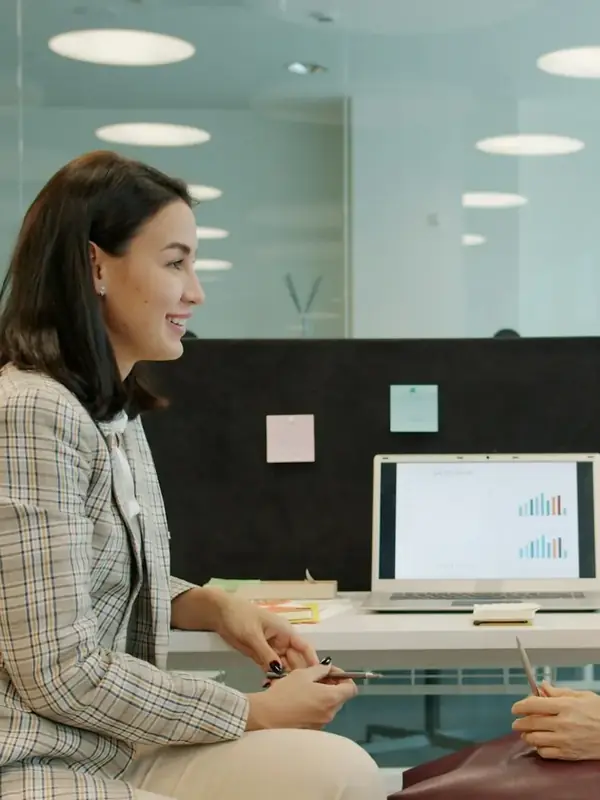Zwei Frauen im Gespräch in einem hellen Büro, ein Computer zwischen ihnen.